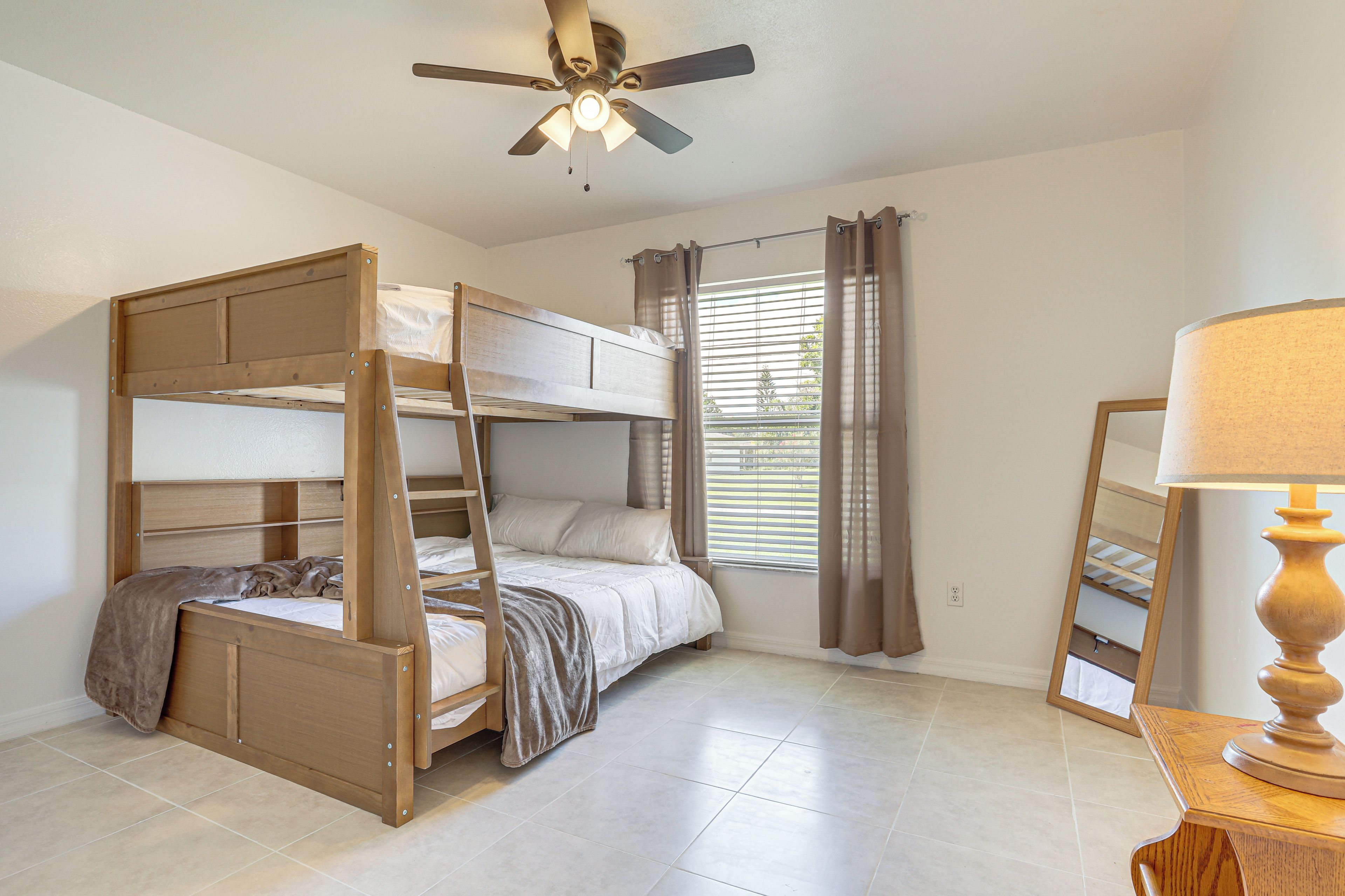 Une chambre avec un lit superposé, un ventilateur de plafond, une fenêtre avec des stores et une table de chevet avec une lampe.