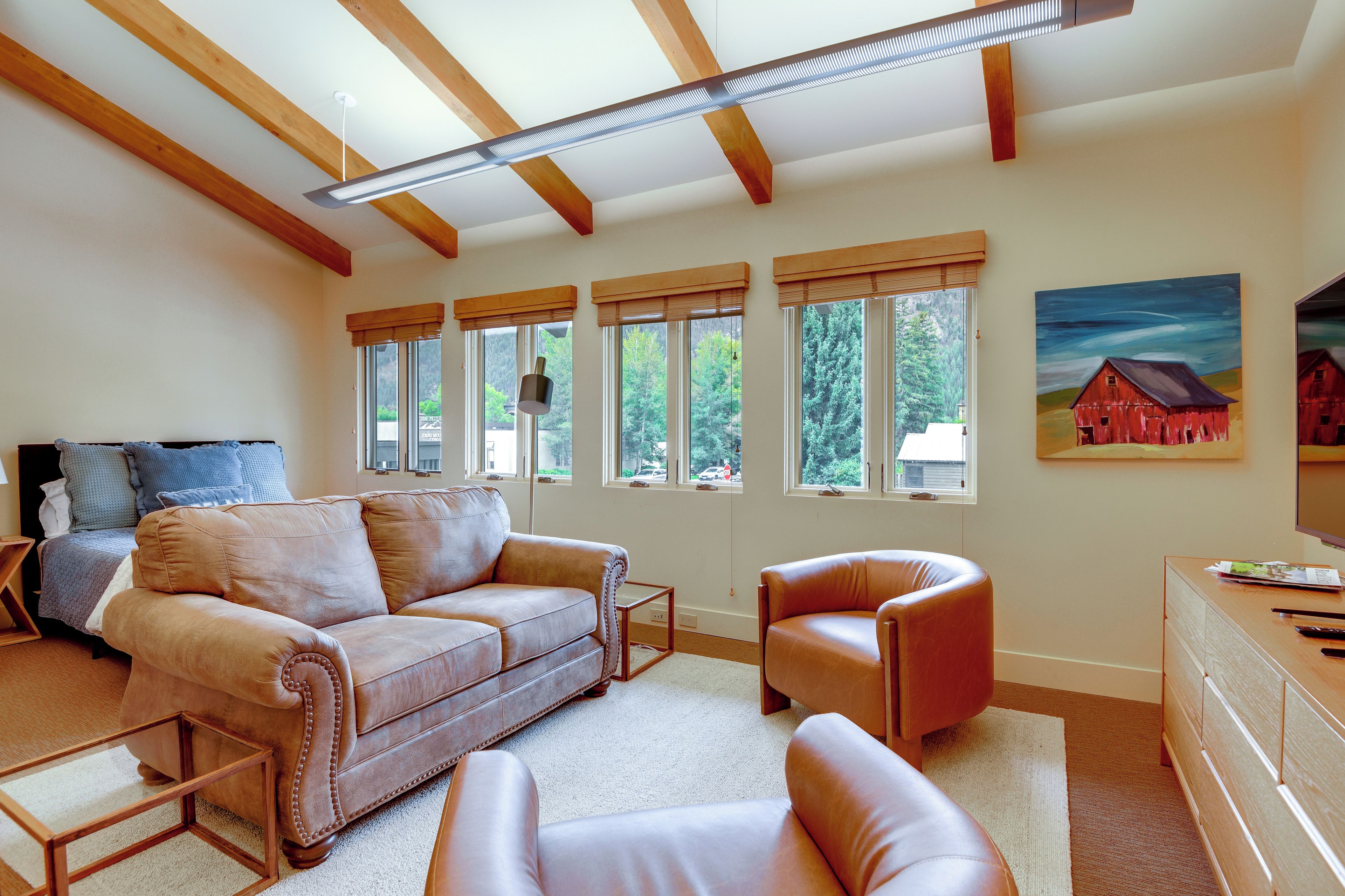 Ein Wohnzimmer mit einem braunen Ledersofa, einem Holztisch und einem roten Sessel. Große Fenster mit Holzrahmen bieten Blick auf Bäume und ein Haus.