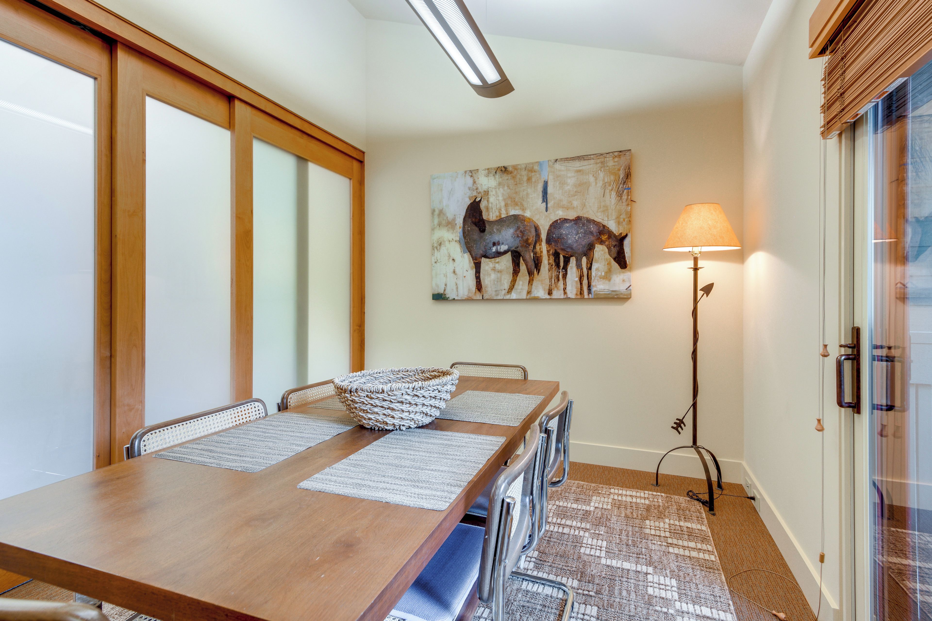 A dining area with a wooden table, chairs, a woven basket, and a painting of horses on the wall.