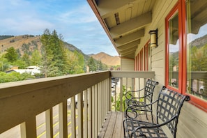 A wooden balcony with two metal chairs, a view of a parking lot, and a mountain in the background.