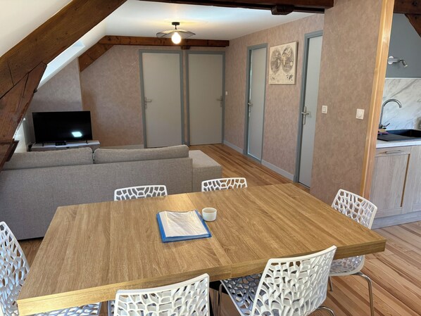 A cozy room with a wooden dining table, white chairs, a sofa, and a television.