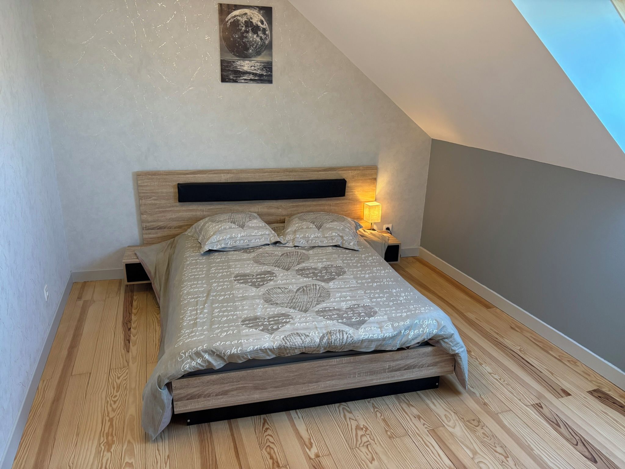 Una camera da letto con soffitto spiovente, un letto con biancheria a motivi, un comodino con una lampada e un quadro appeso al muro.
