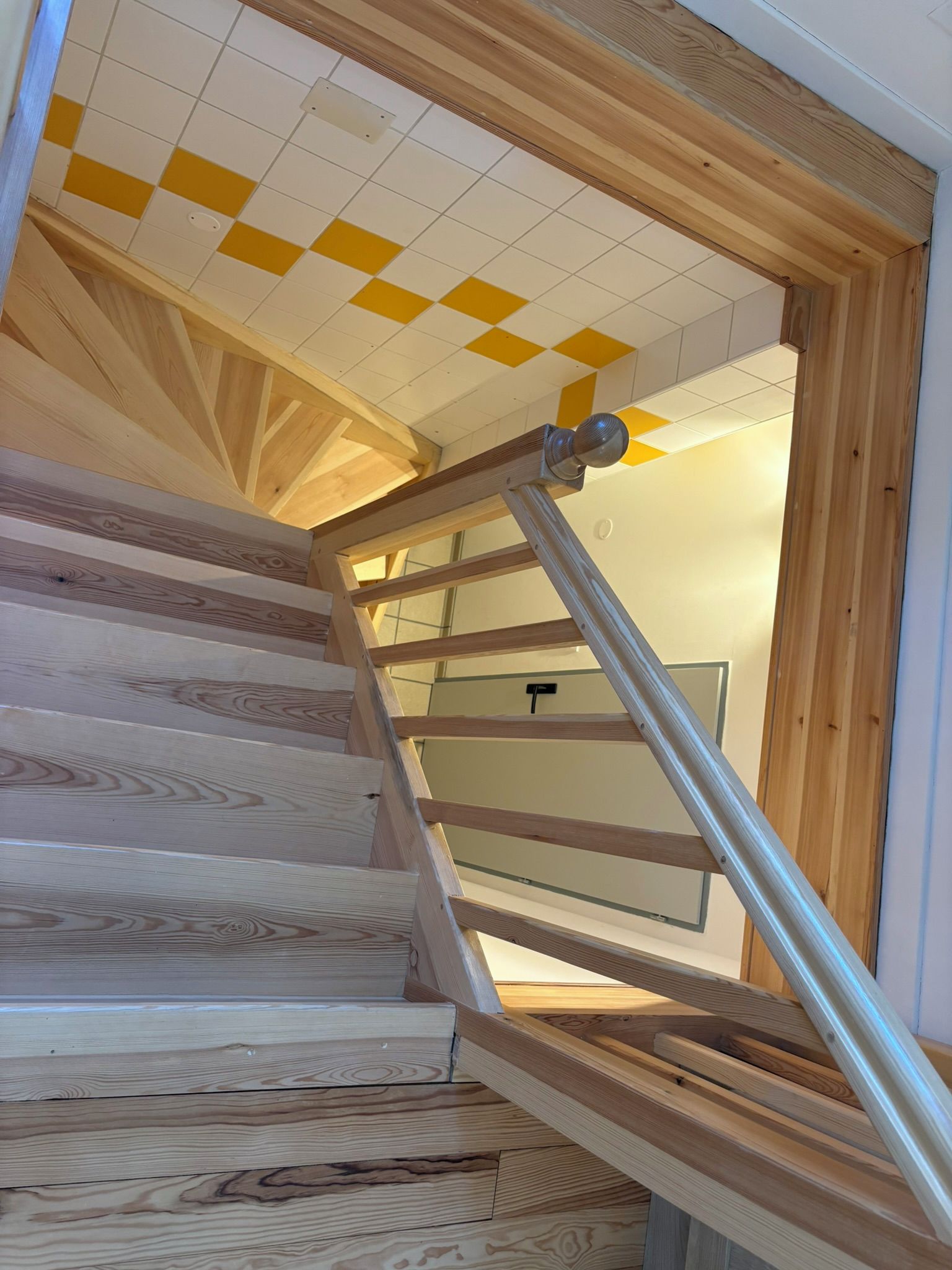 Een houten trap met een metalen leuning die leidt naar een ruimte met een wit plafond en gele tegels.