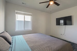 Un dormitorio con una cama, ventilador de techo, ventana con persianas y televisor de pantalla plana montado en la pared.
