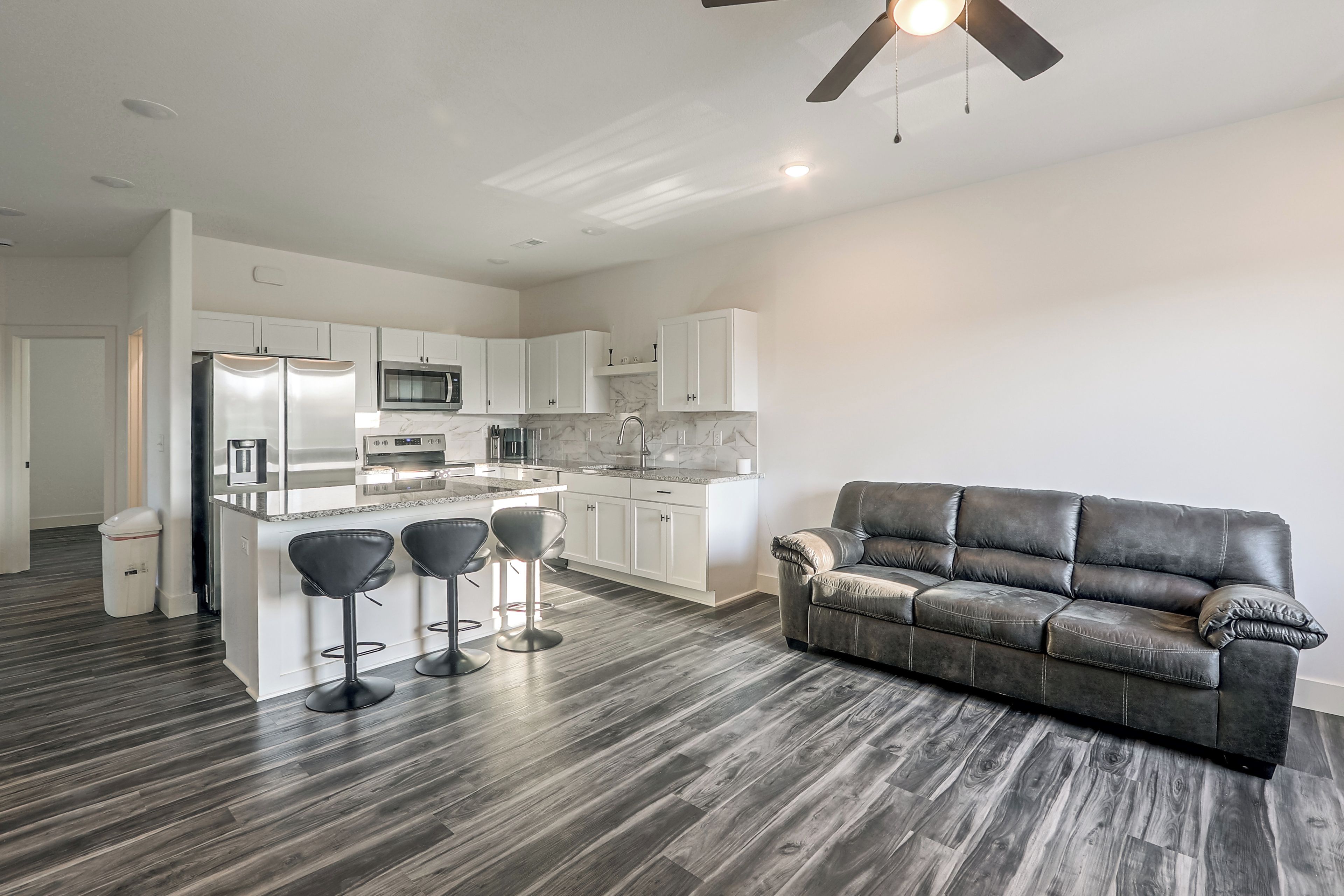A modern kitchen with white cabinets, a central island with bar stools, a dark leather sofa, and a ceiling fan.