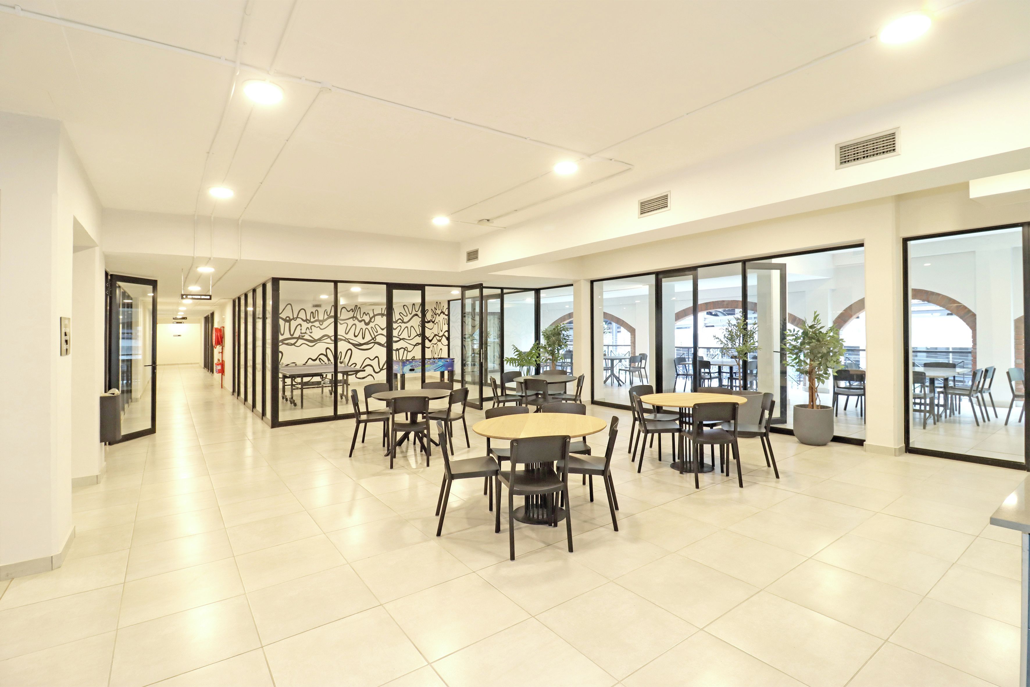 A modern indoor space with multiple tables and chairs, large windows, and a clean, tiled floor.