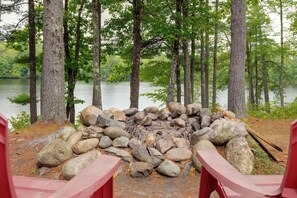 Un camping con un brasero rodeado de rocas, sillas rojas y un lago al fondo.
