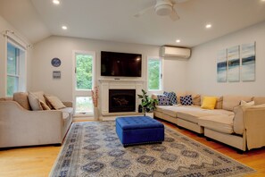 Una sala de estar con chimenea, dos sofás, un reposapiés azul y un televisor de pantalla plana.