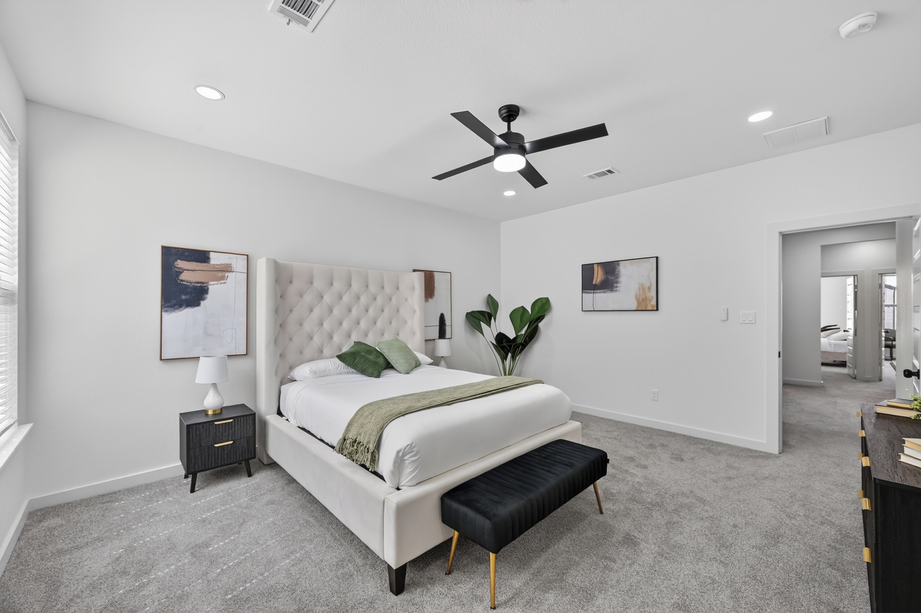 A bedroom with a tufted headboard, a bed with white bedding, a black bench, a nightstand with a lamp, and two wall-mounted paintings.