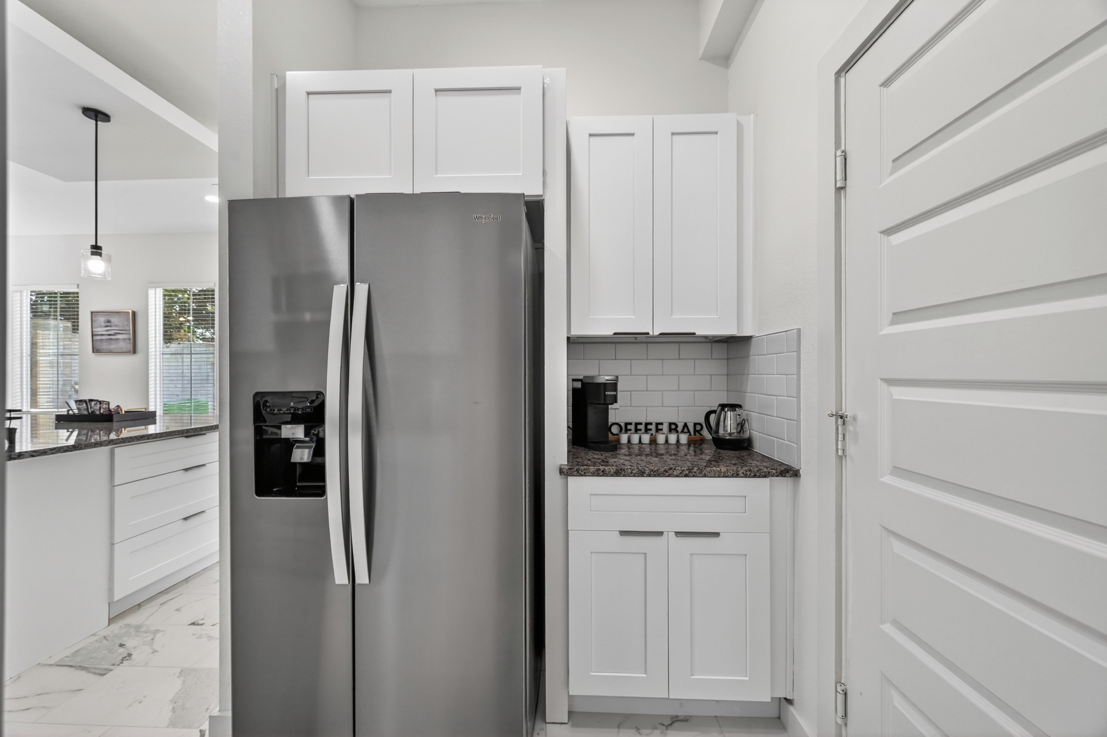 A modern kitchen with white cabinetry, a stainless steel refrigerator, a coffee station, and a marble floor.