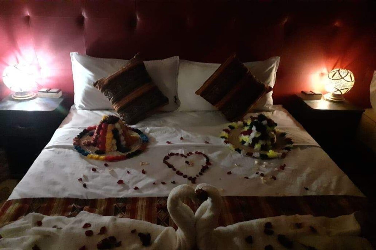 A bed with a heart-shaped decoration made of rose petals and a swan-shaped decoration, a red upholstered headboard, and two bedside tables with lamps.