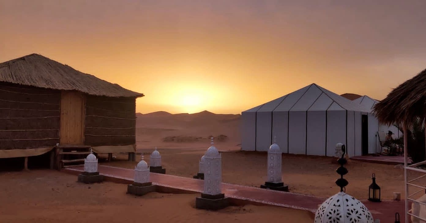 A desert landscape with a thatched-roof building and a modern structure with a peaked roof.