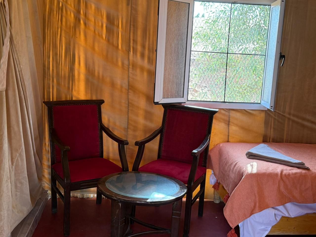 Habitació amb llit, dues cadires, taula rodona petita i finestra amb vista a vegetació.