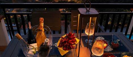 Vista al atardecer desde un balcón con una mesa preparada para una comida, que incluye vino y aperitivos.