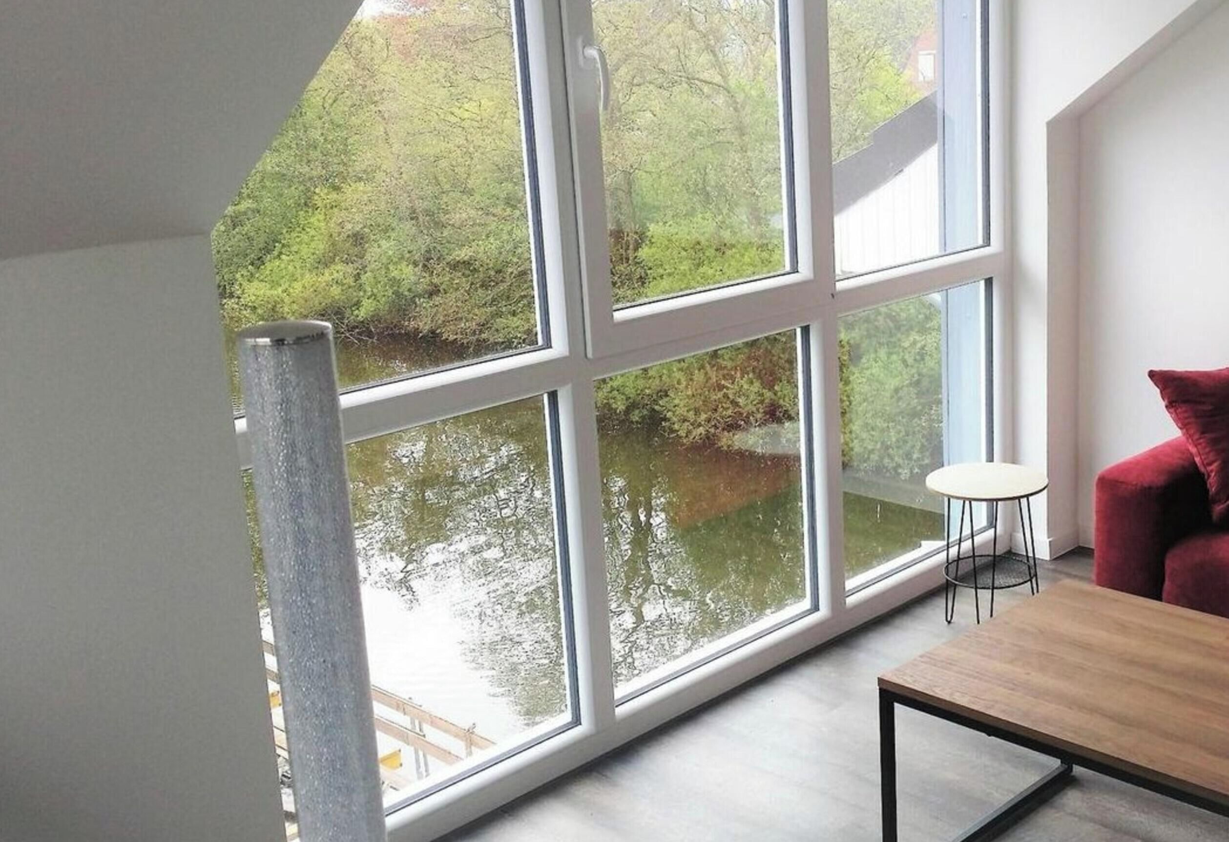 A room with a large window overlooking a body of water and greenery. There is a wooden table and a red chair in the room.