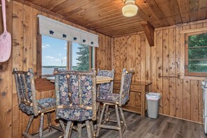 Innenansicht einer Holzhütte mit Esstisch und Stühlen, einem Fenster mit Blick und einem Deckenventilator.