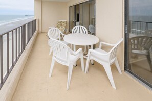 Ein Balkon mit einem runden Tisch und sechs weißen Stühlen, mit Blick auf einen Strand mit einem Steg.