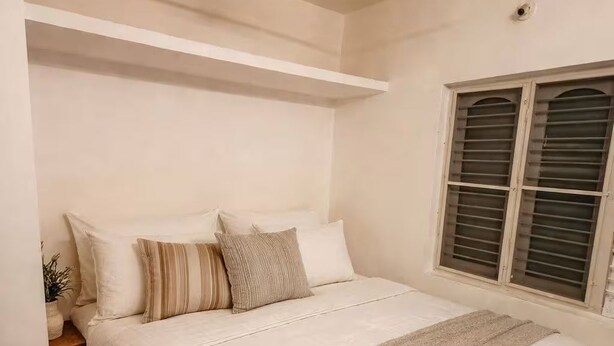 Un dormitorio con cama, ventilador de techo, ventana con persianas y mesita de noche con una planta.