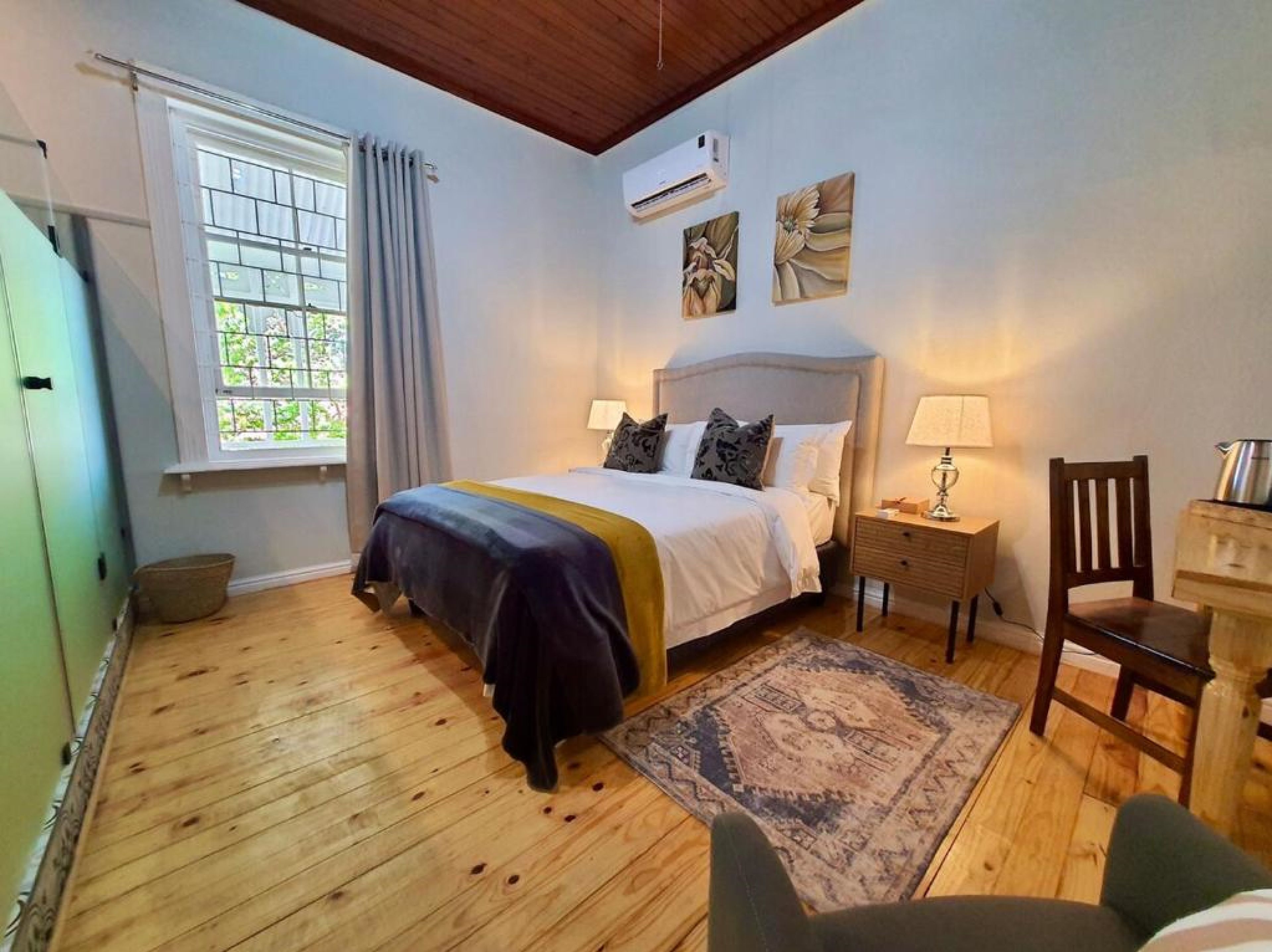 A bedroom with a bed, a nightstand, a chair, a window with curtains, and a wall-mounted air conditioner.