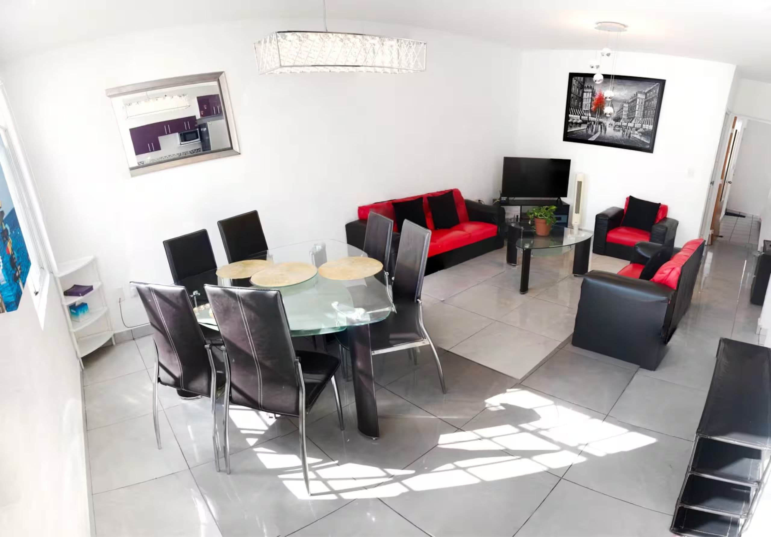 Ein modernes Wohnzimmer mit Essbereich, ausgestattet mit einem Glastisch, schwarzen Stühlen, einem roten Sofa und einem Flachbildfernseher.