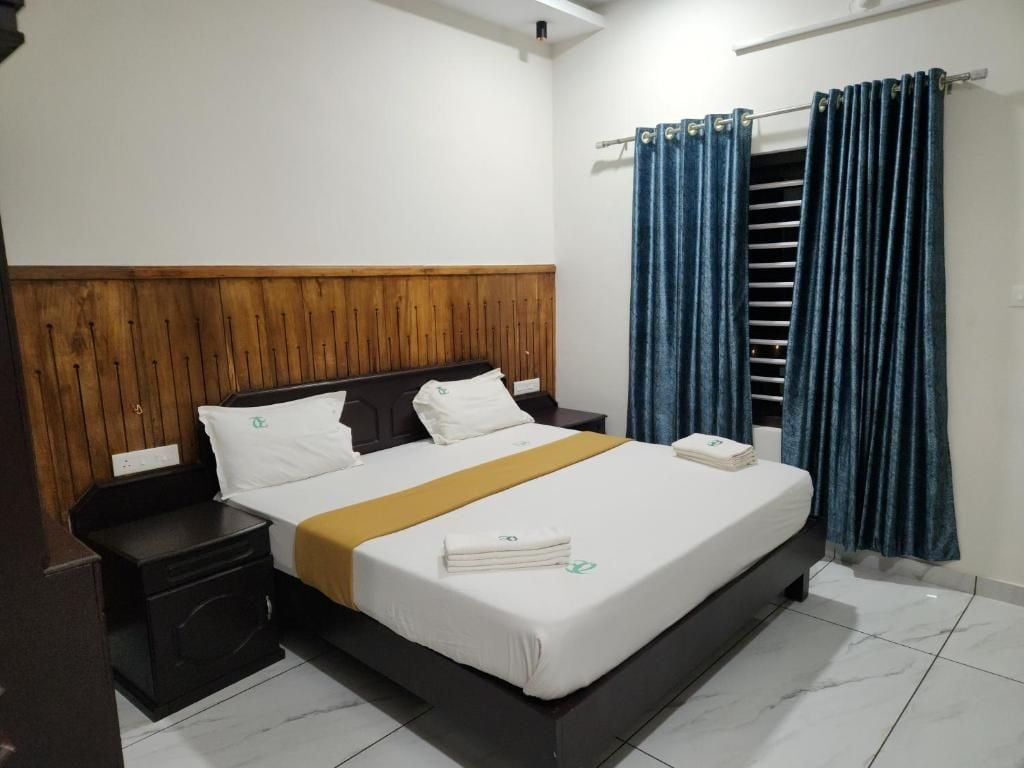 Habitación de hotel con cabecera de madera, cama con ropa blanca, mesita de noche y cortina azul.