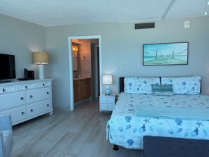 A bedroom with a bed, a television, a dresser, and a bathroom visible through an open door. - Peck7NE Oceanfront Condo - Magical Views, Heated Pool, Hot Tub, Beach Decor (Daytona Beach)