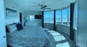 Un dormitorio con una cama grande, ventilador de techo, televisión y vista al océano.