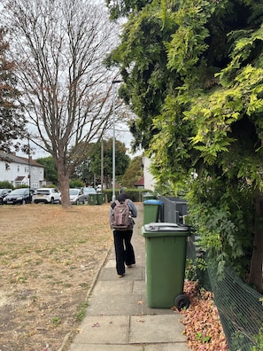 A sidewalk with a person walking away from the camera, a tree, parked cars, and green bins. - Studio Sleeps 2 With Garden and Parking (London)