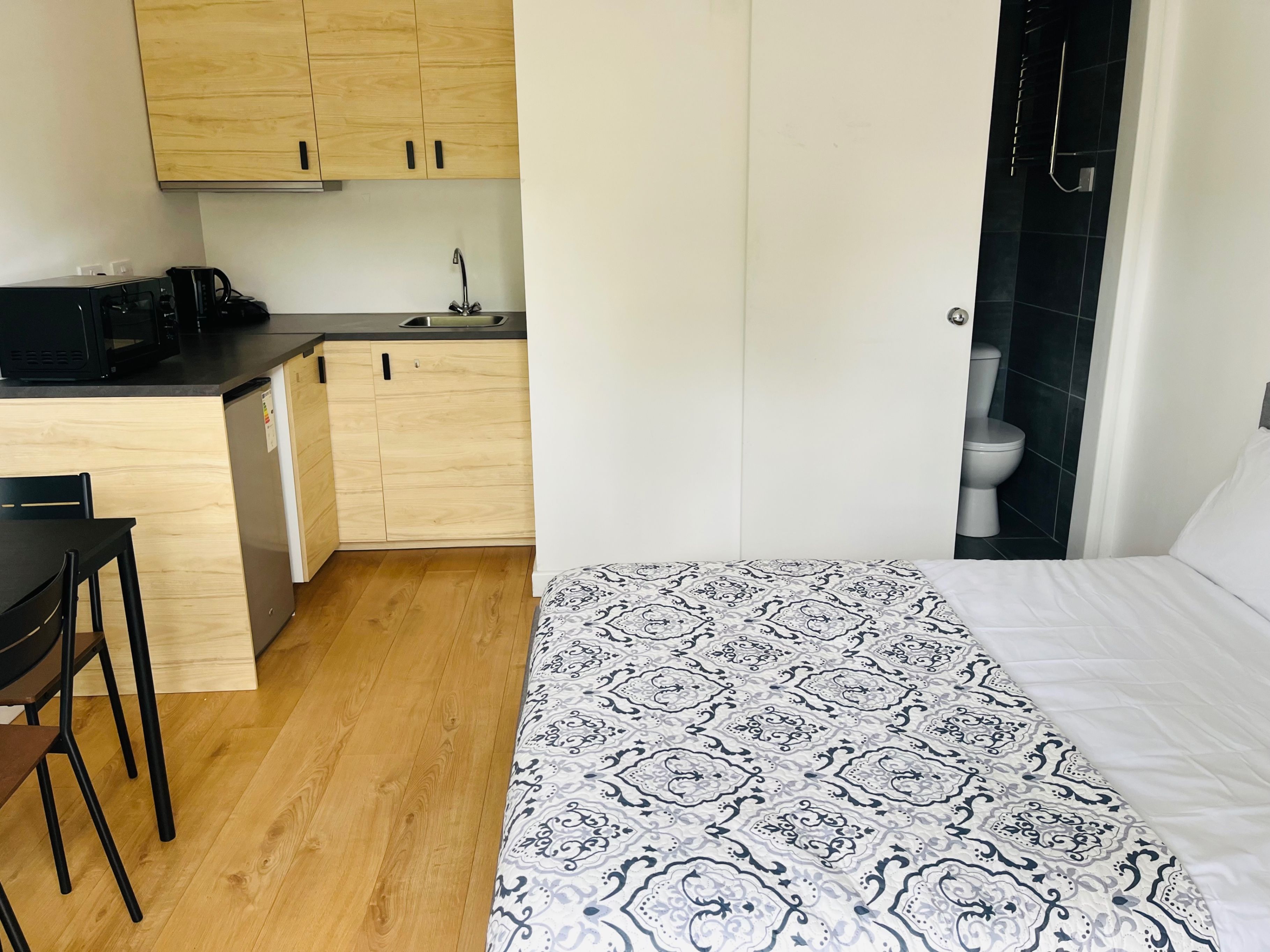 A compact room with a kitchenette, a bed with patterned bedding, and a toilet in the adjacent bathroom.