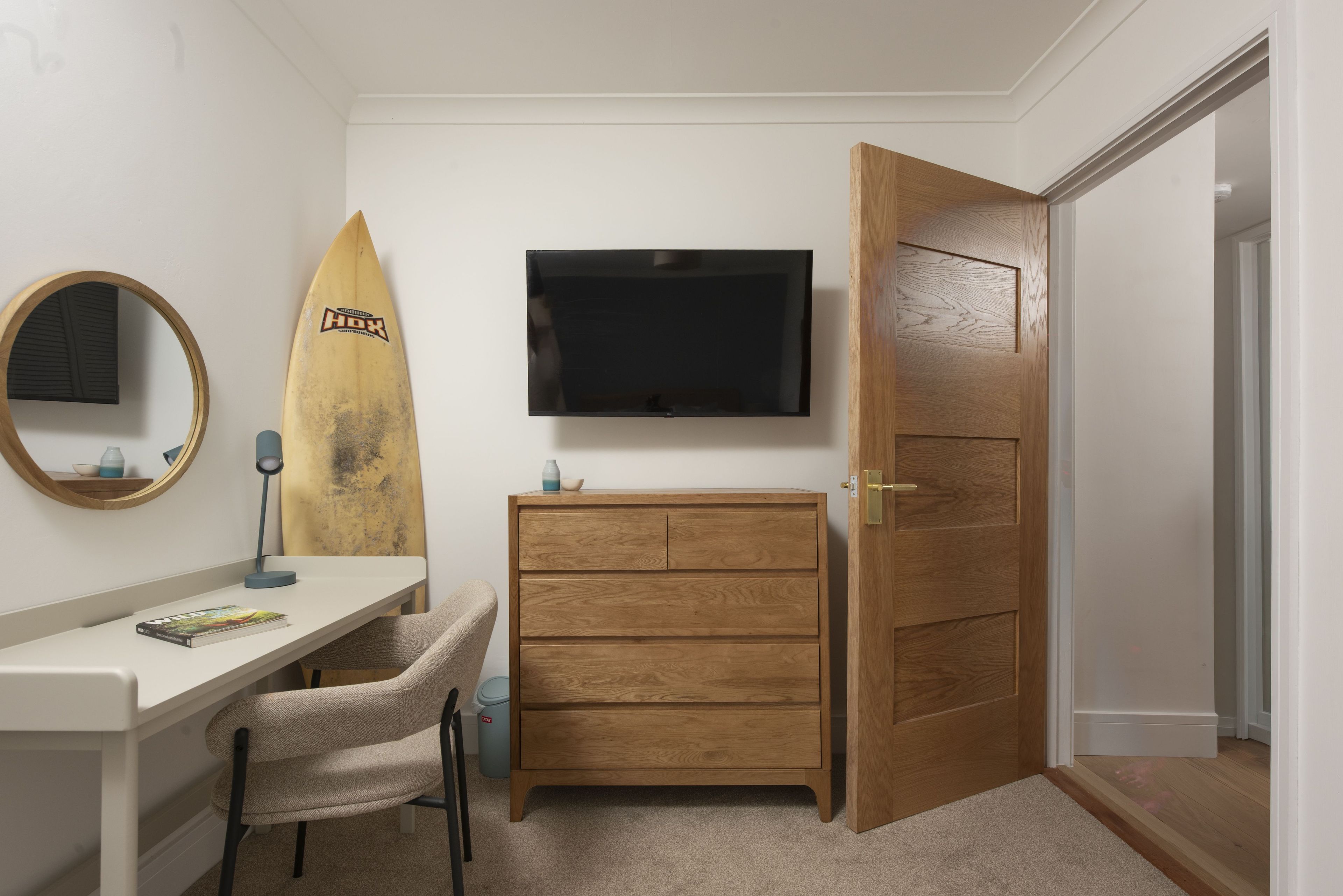 Ein Schlafzimmer mit einem Holzkasten, einem Schreibtisch mit Stuhl, einem Surfbrett, einem Fernseher und einer Tür.