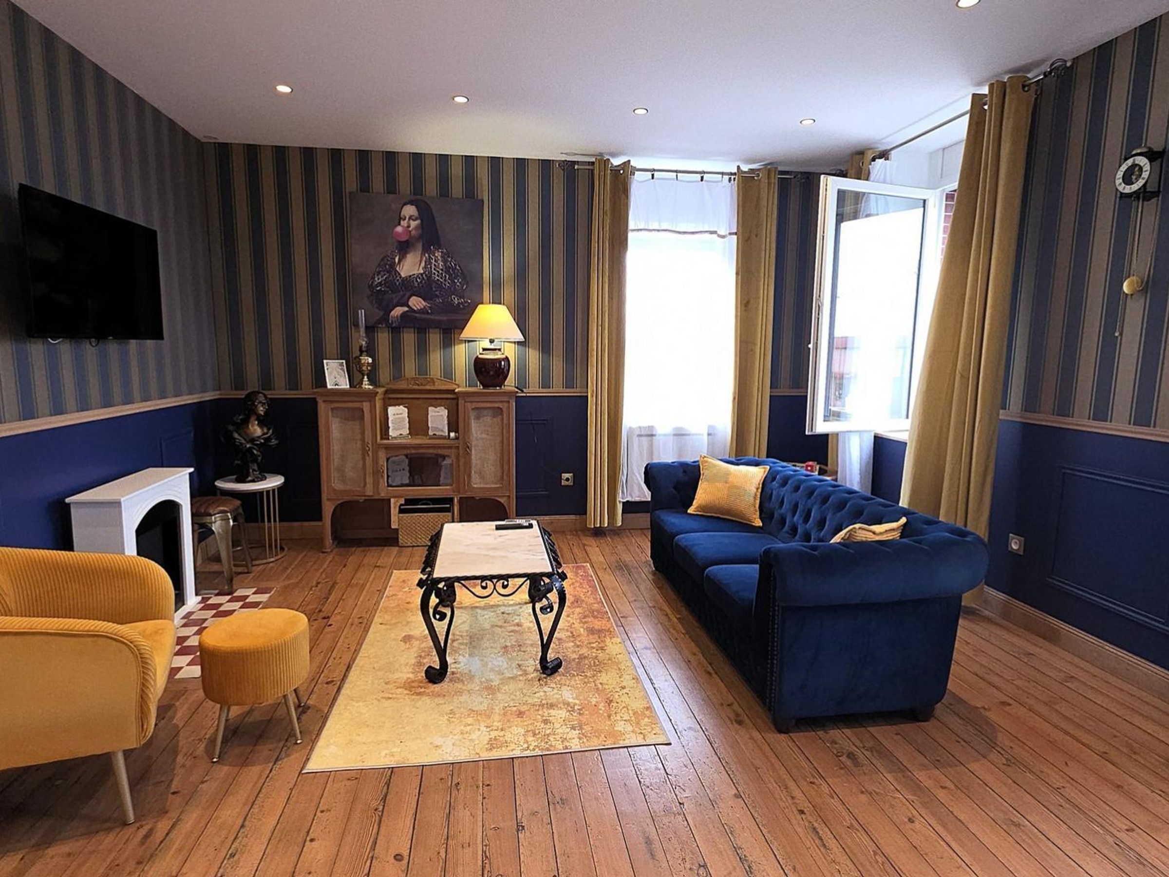 Een woonkamer met een blauwe bank, een gele fauteuil, een salontafel en een televisie aan de muur.
