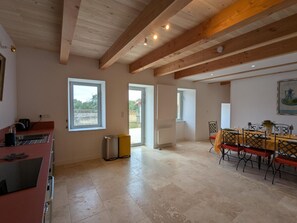Habitació àmplia amb zona de cuina, taula de menjador i cadiretes. Hi ha finestres que deixen entrar llum natural i ofereixen vistes a l'exterior.