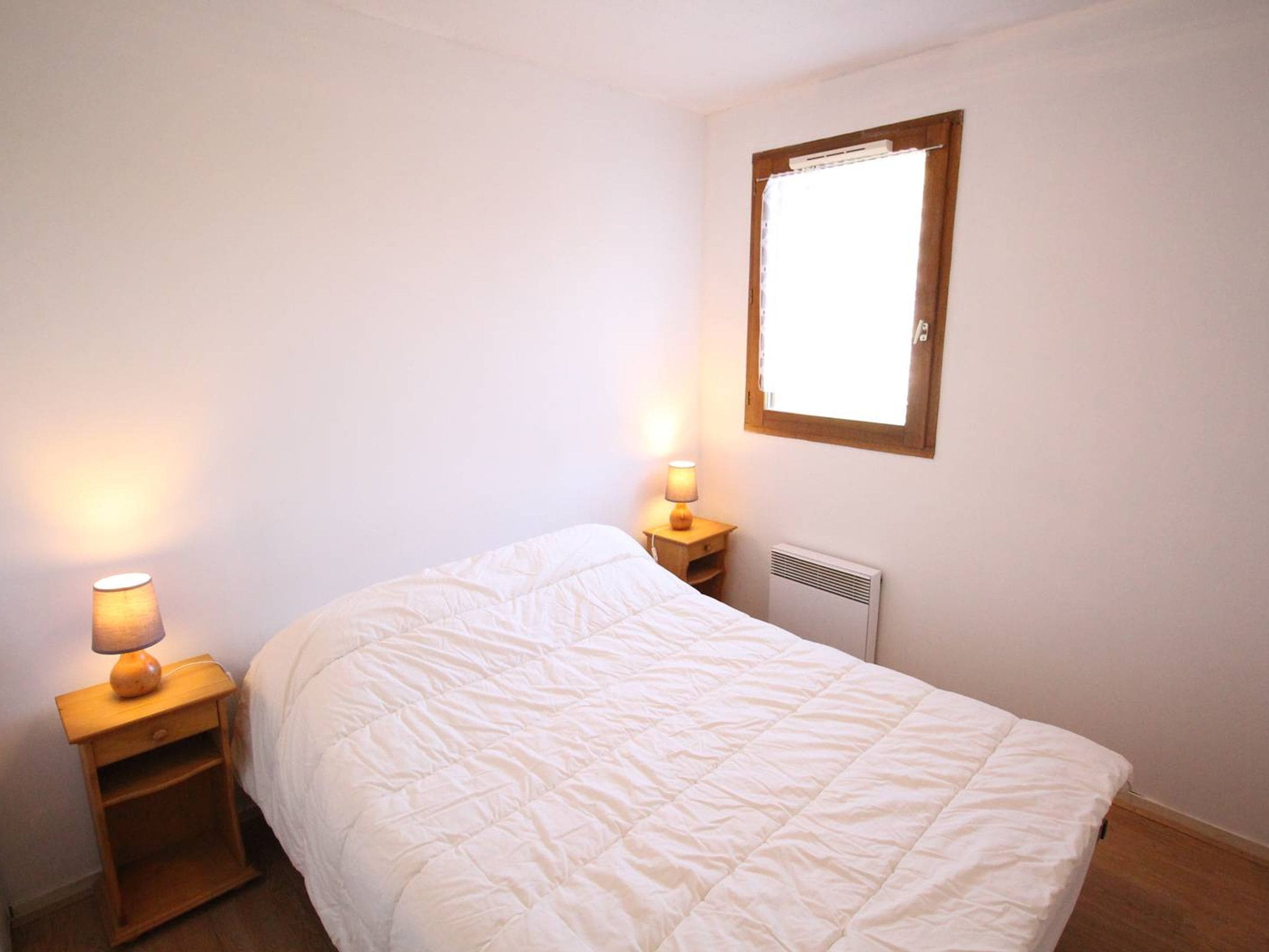 A single bed with a white duvet, a bedside table with a lamp, and a window with a wooden frame.