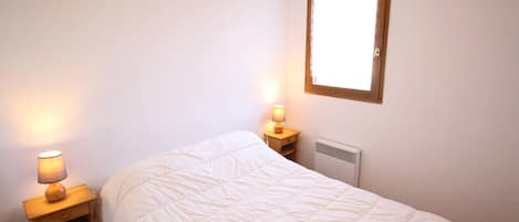 Un lit simple avec une couette blanche, une table de chevet avec une lampe et une fenêtre avec un cadre en bois.