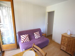 Ein kleines Wohnzimmer mit einem violetten Sofa, zwei gemusterten Kissen, einem Holztisch mit Teller und Messer sowie einer Holzkabine mit Schubladen.