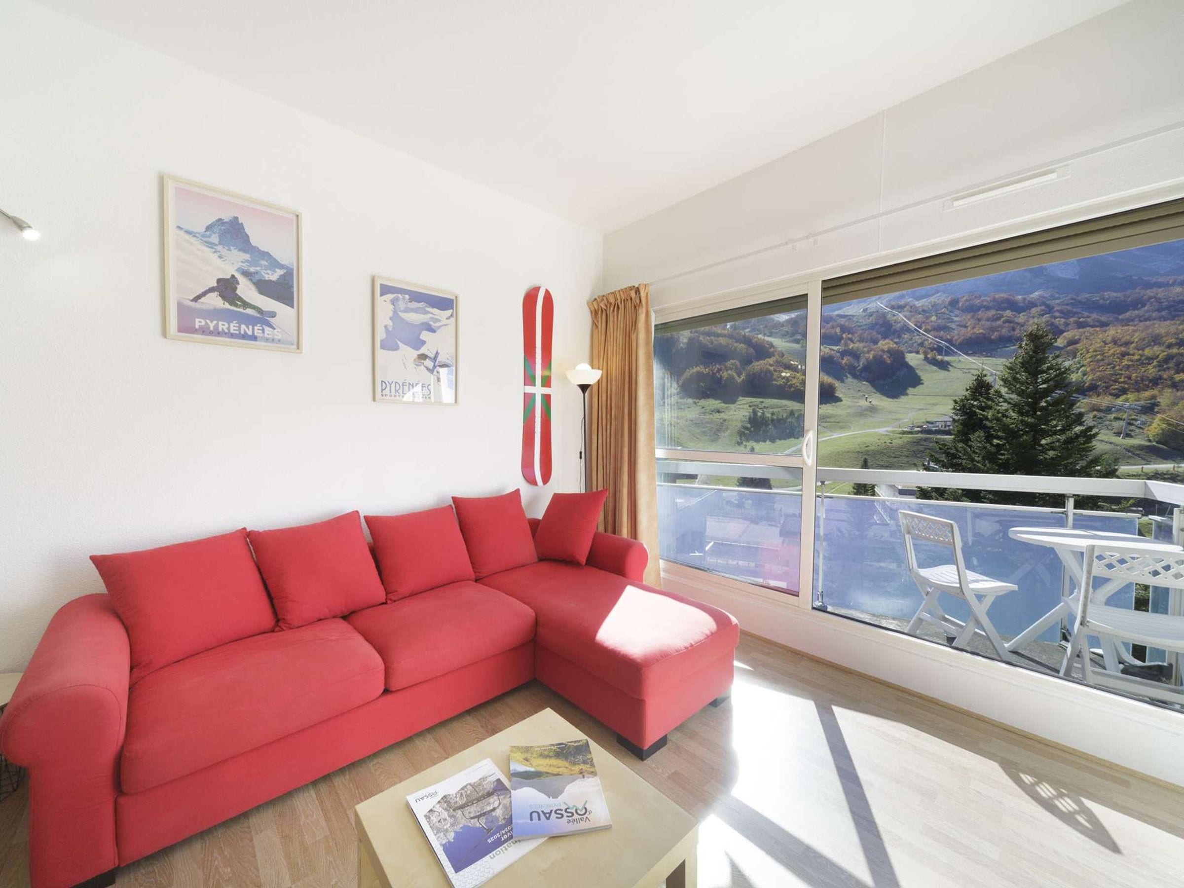 A modern living room with a red sofa, a small table with magazines, and a large window offering a view of a mountainous landscape.