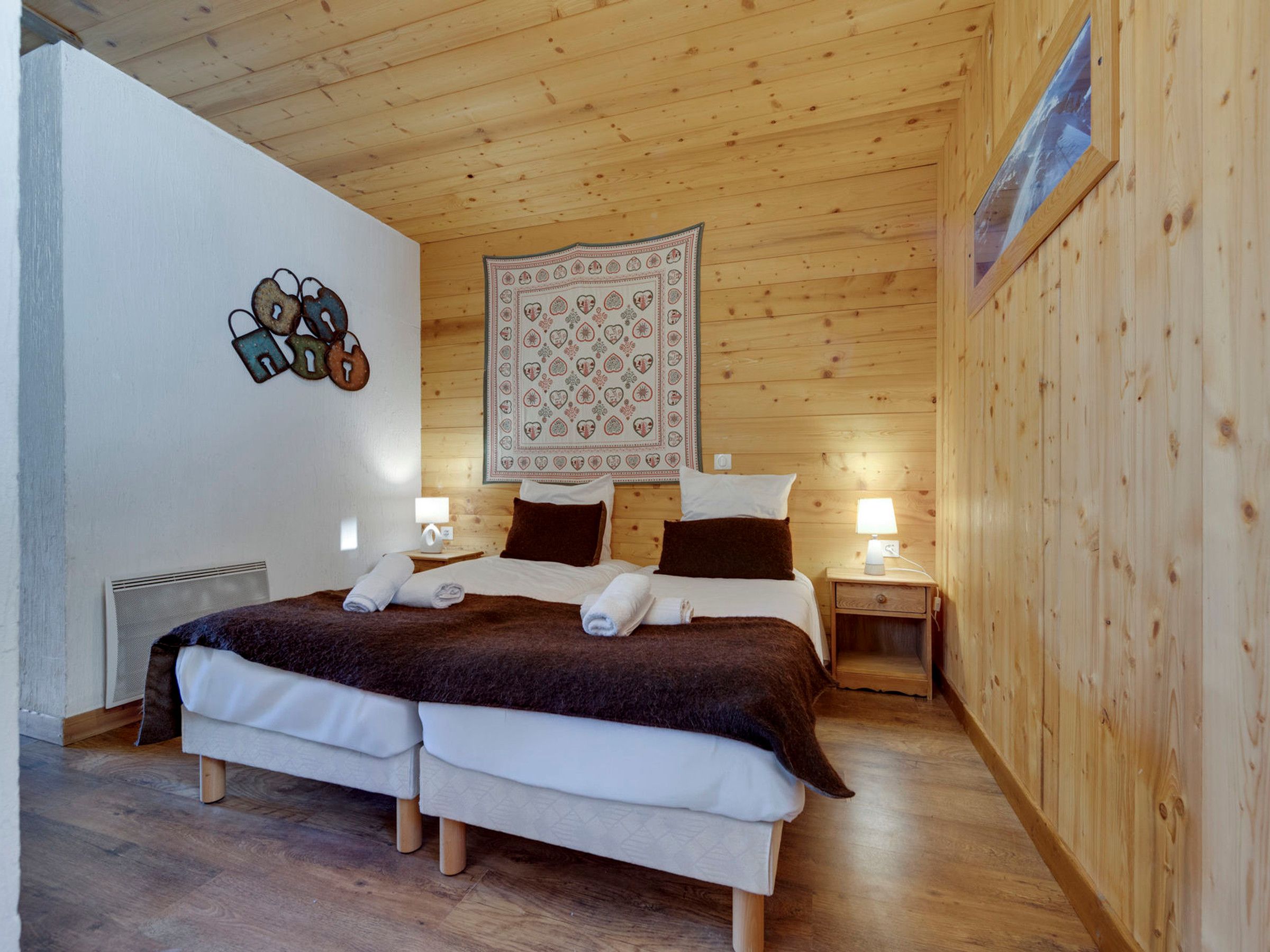 Uma cama de casal com cobertor marrom, duas mesas de cabeceira com luminárias, tapeçaria na parede e teto de madeira.