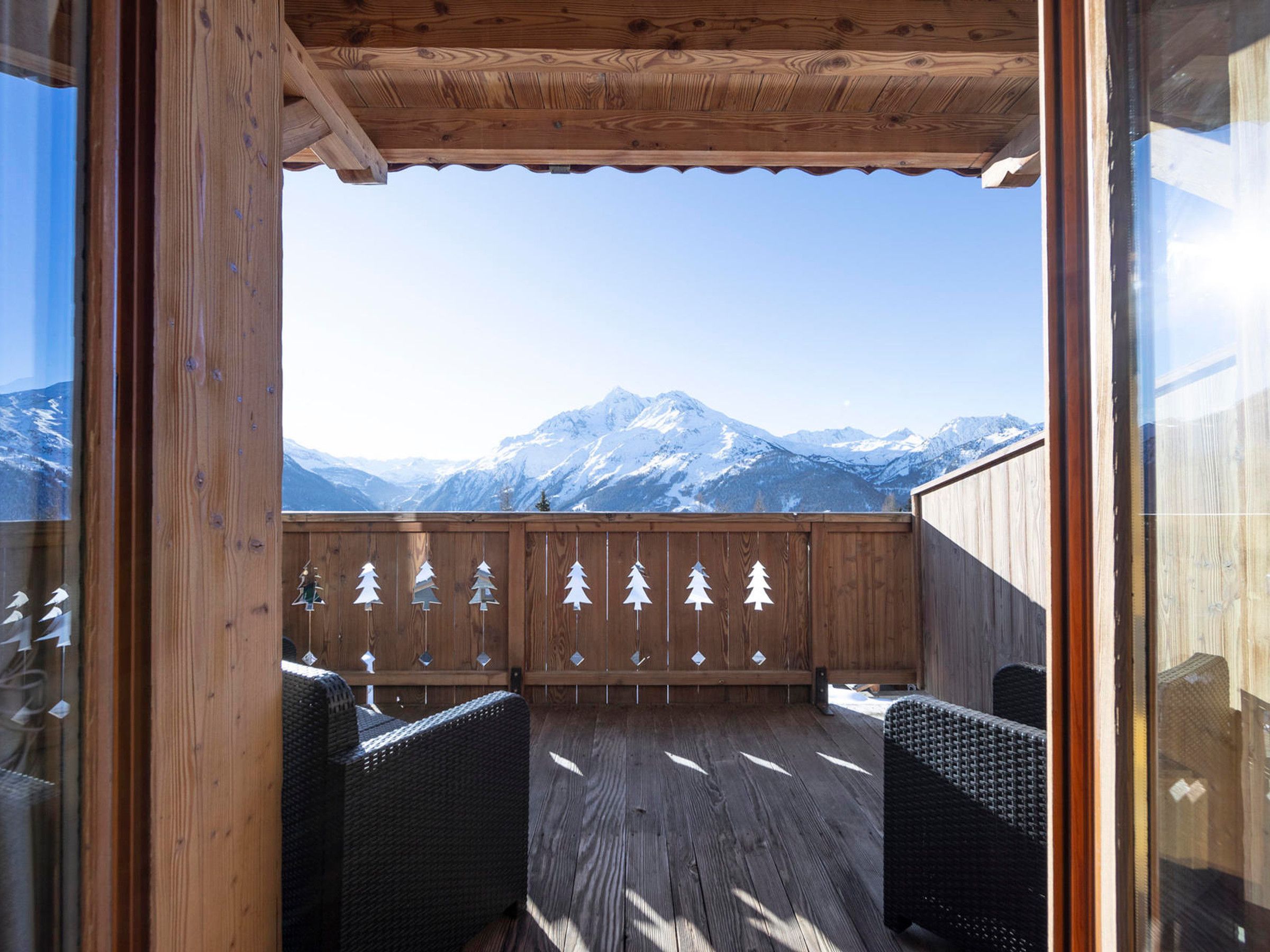 Une terrasse en bois avec des chaises en osier, une vue sur des montagnes enneigées et une balustrade en bois décorée de motifs végétaux.