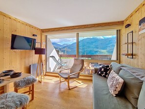 A cozy room with a wooden interior, a flat-screen TV mounted on the wall, a sofa with a floral pillow, and a dining area with a table and chairs. - Cozy ski-in/ski-out apartment, south-facing balcony (Les Menuires)