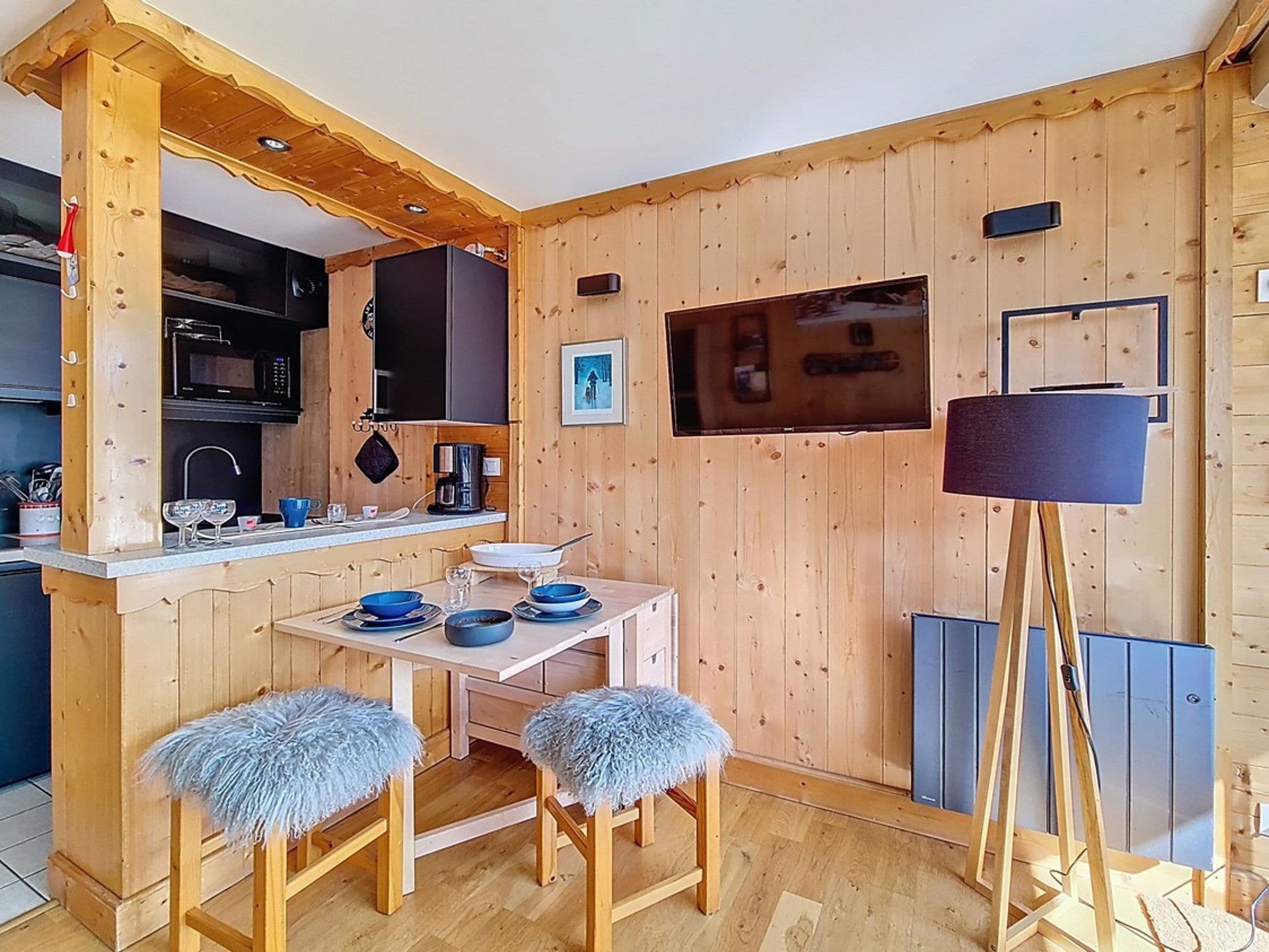 A compact wooden room with a kitchen area, a dining table with blue tableware, and a floor lamp.