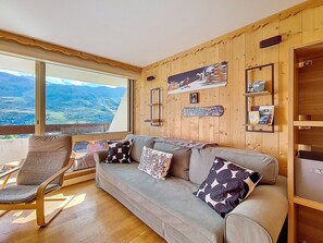 A cozy room with a large window, a sofa with patterned cushions, and a wooden wall with various decorations. - Cozy ski-in/ski-out apartment, south-facing balcony (Les Menuires)