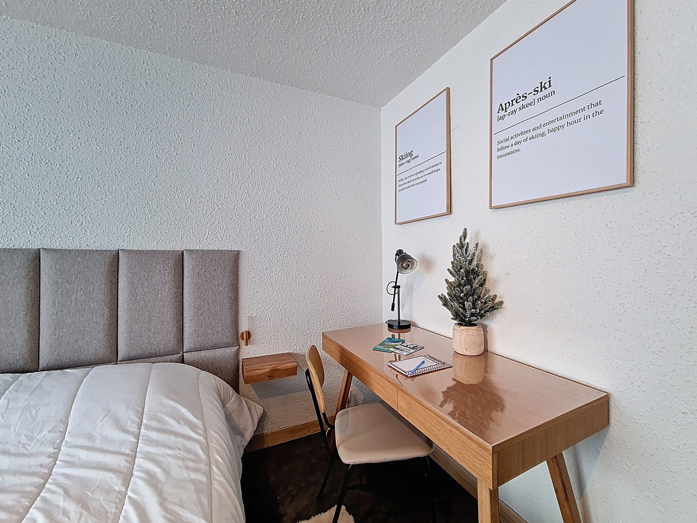 A bedroom with a bed, a desk, a chair, a small plant, and two framed informational posters on the wall.