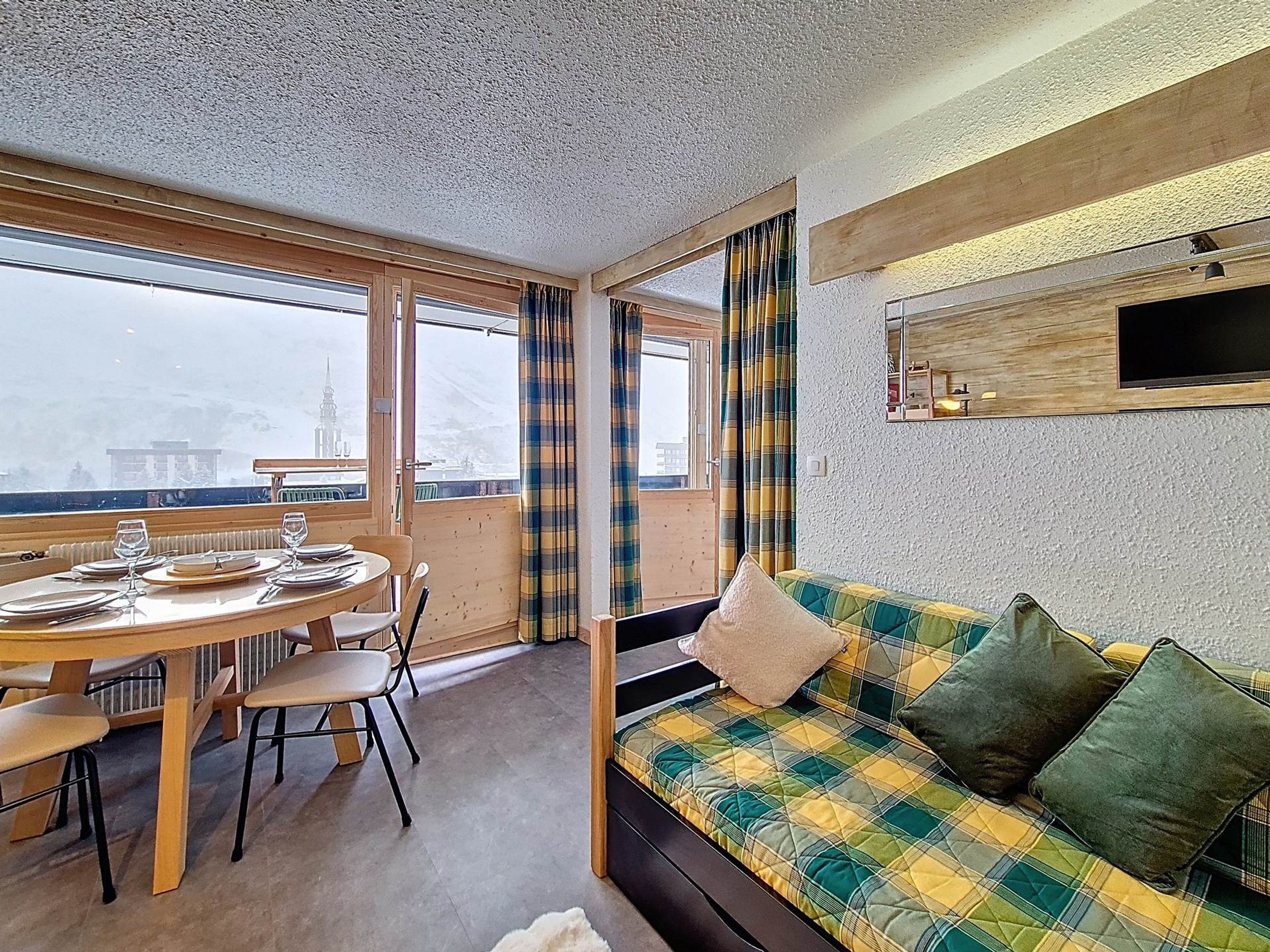 Ein Zimmer mit Esstisch für vier Personen, einer Couch mit kariertem Bezug und einem großen Fenster mit Vorhängen.