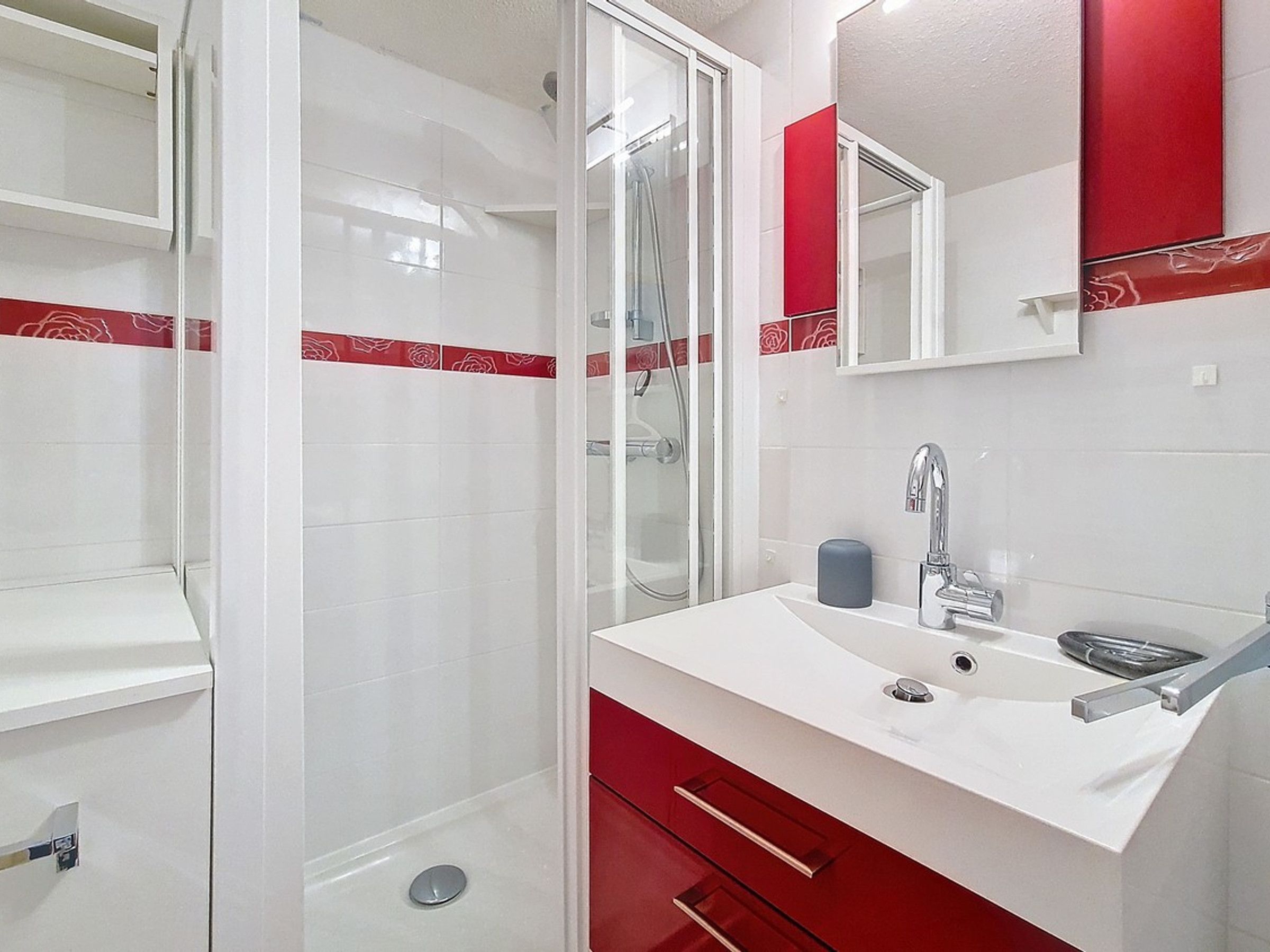 A modern bathroom with a glass shower enclosure, a white sink with a red cabinet, and a mirror above the sink.
