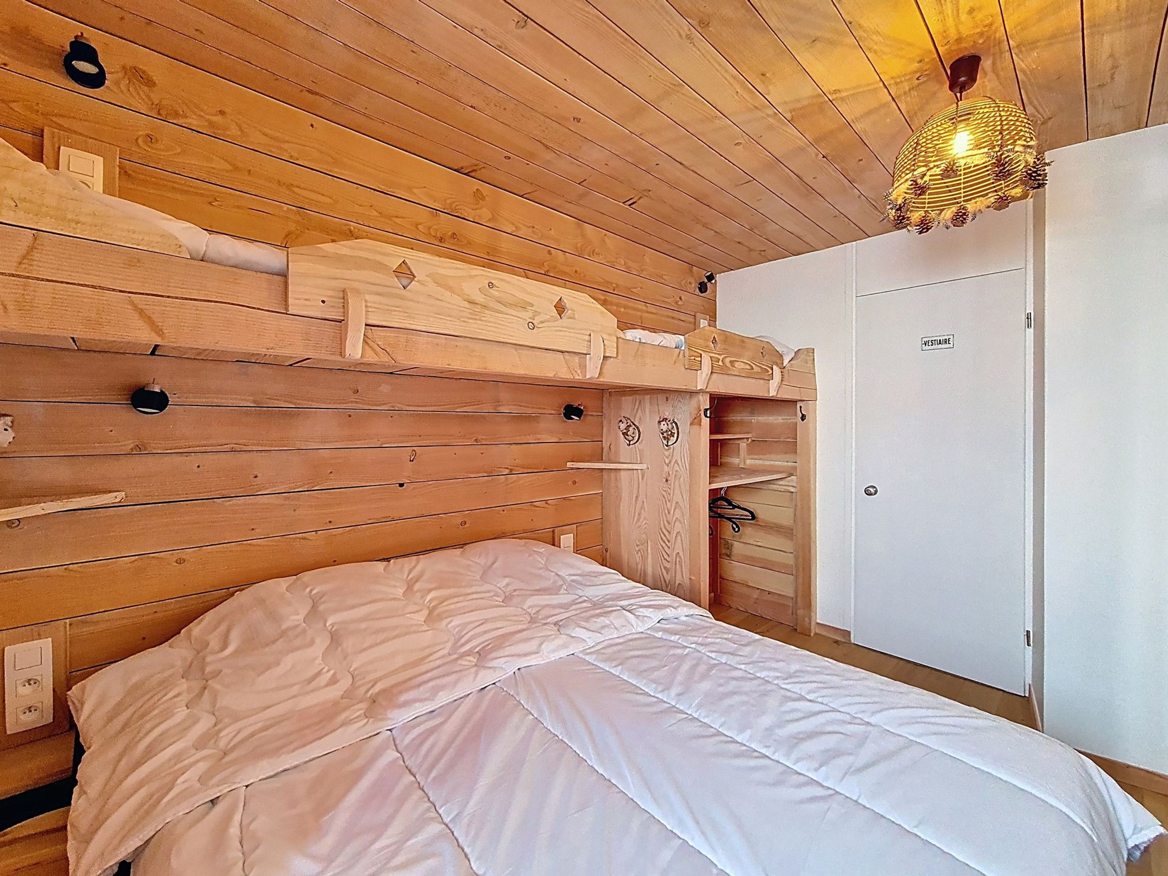A wooden bunk bed with a white mattress, a wooden wardrobe, and a hanging light fixture.