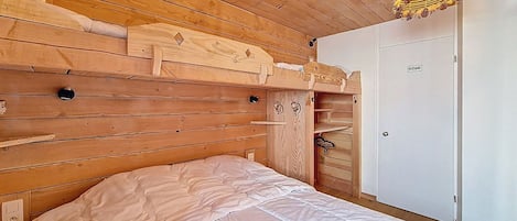 A wooden bunk bed with a white mattress, a wooden wardrobe, and a hanging light fixture.