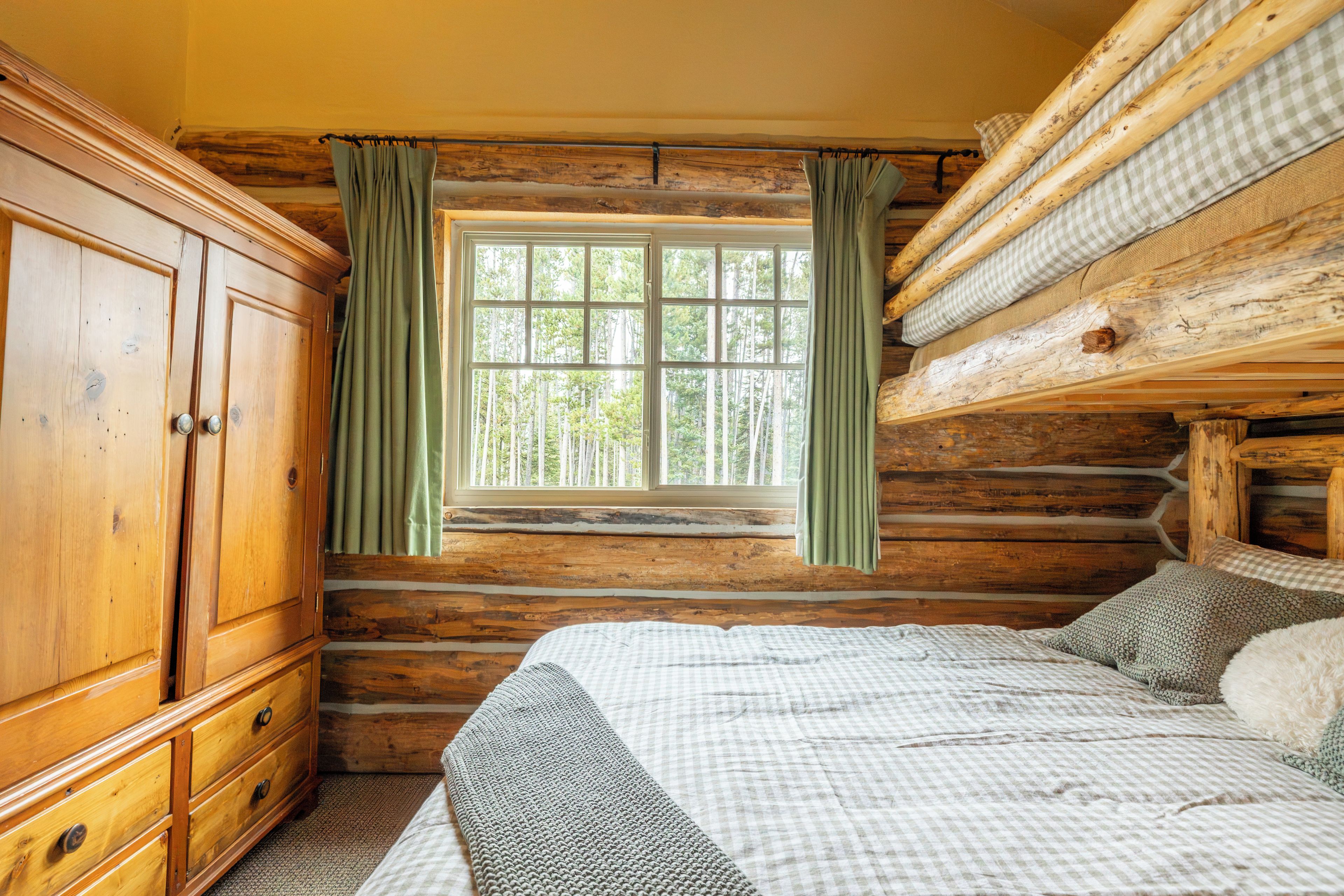 A log cabin room with a large wooden wardrobe, a bed with a checkered bedspread, and a window with curtains.
