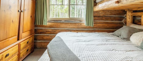 A log cabin room with a large wooden wardrobe, a bed with a checkered bedspread, and a window with curtains.