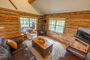 A log cabin interior with a fireplace, sofa, television, and windows. - NEW Stirrup Summit Cabin | Ski-In/Out 2BR Hot Tub (Big Sky)