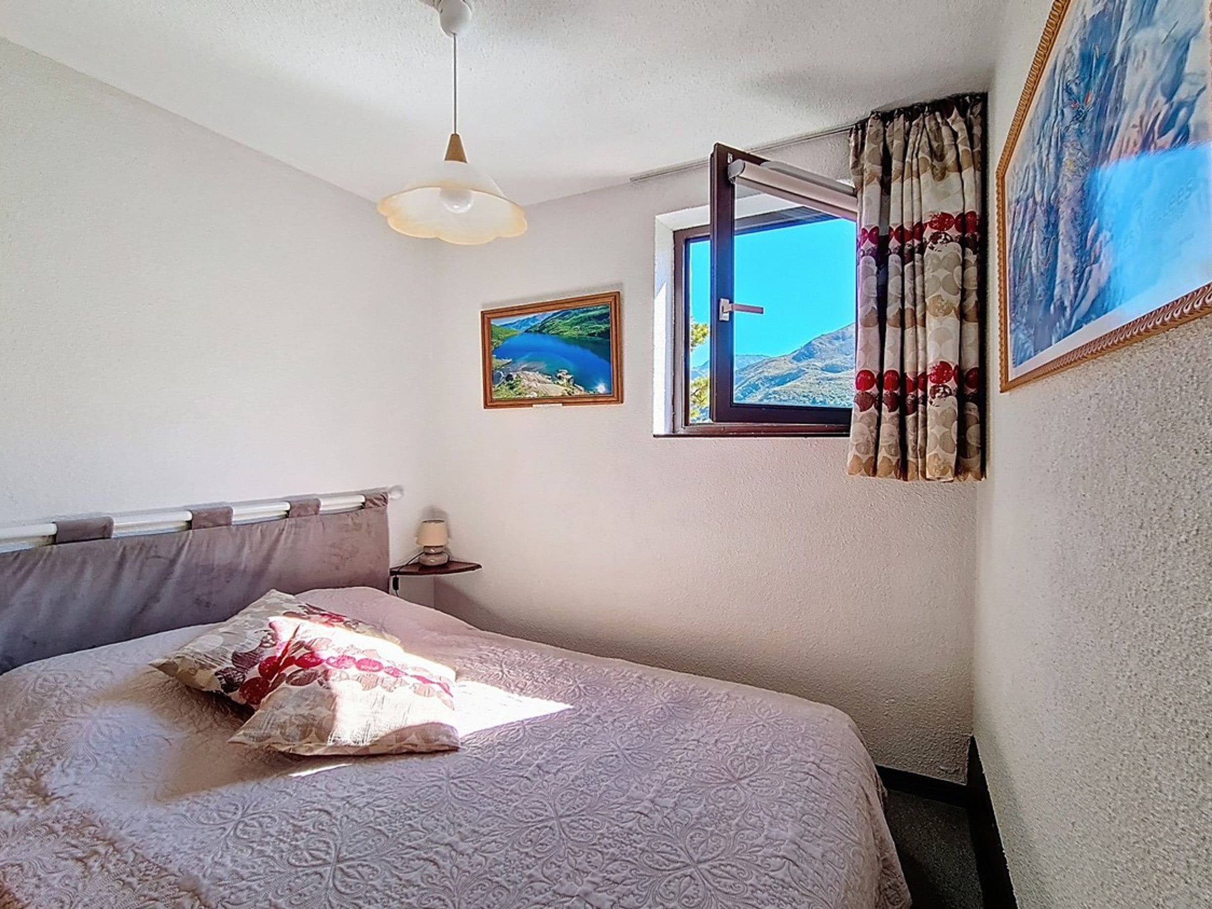 A bedroom with a bed, a window with curtains, a painting, and a ceiling light.